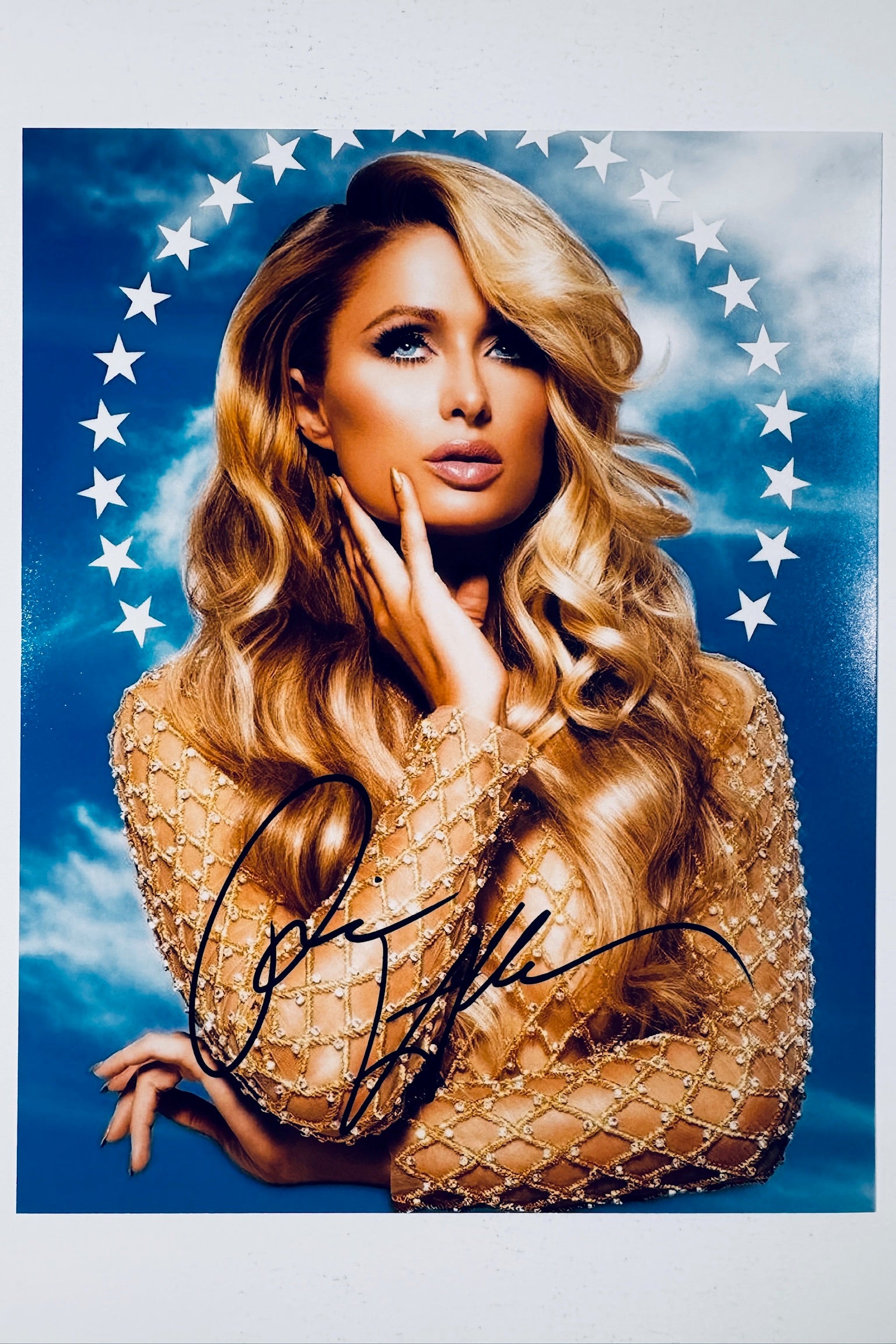 Signed full photo of Paris Hilton posing in front of a blue sky with a ring of white stars, featuring a black autograph.