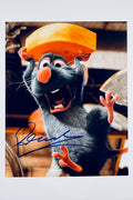 A still from Ratatouille featuring Remy holding a piece of cheese over his head with an excited expression, signed in blue ink on the lower left.
