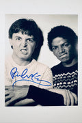 A black-and-white studio portrait of Paul McCartney and Michael Jackson making playful grimaces and pulling funny faces side-by-side, featuring Paul McCartney blue marker signature across the lower portion.