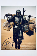 A promotional portrait of Pedro Pascal as the Mandalorian wearing beskar armor and walking through a desert carrying heavy bags and Grogu in a pouch, featuring a black marker signature across the lower foreground.