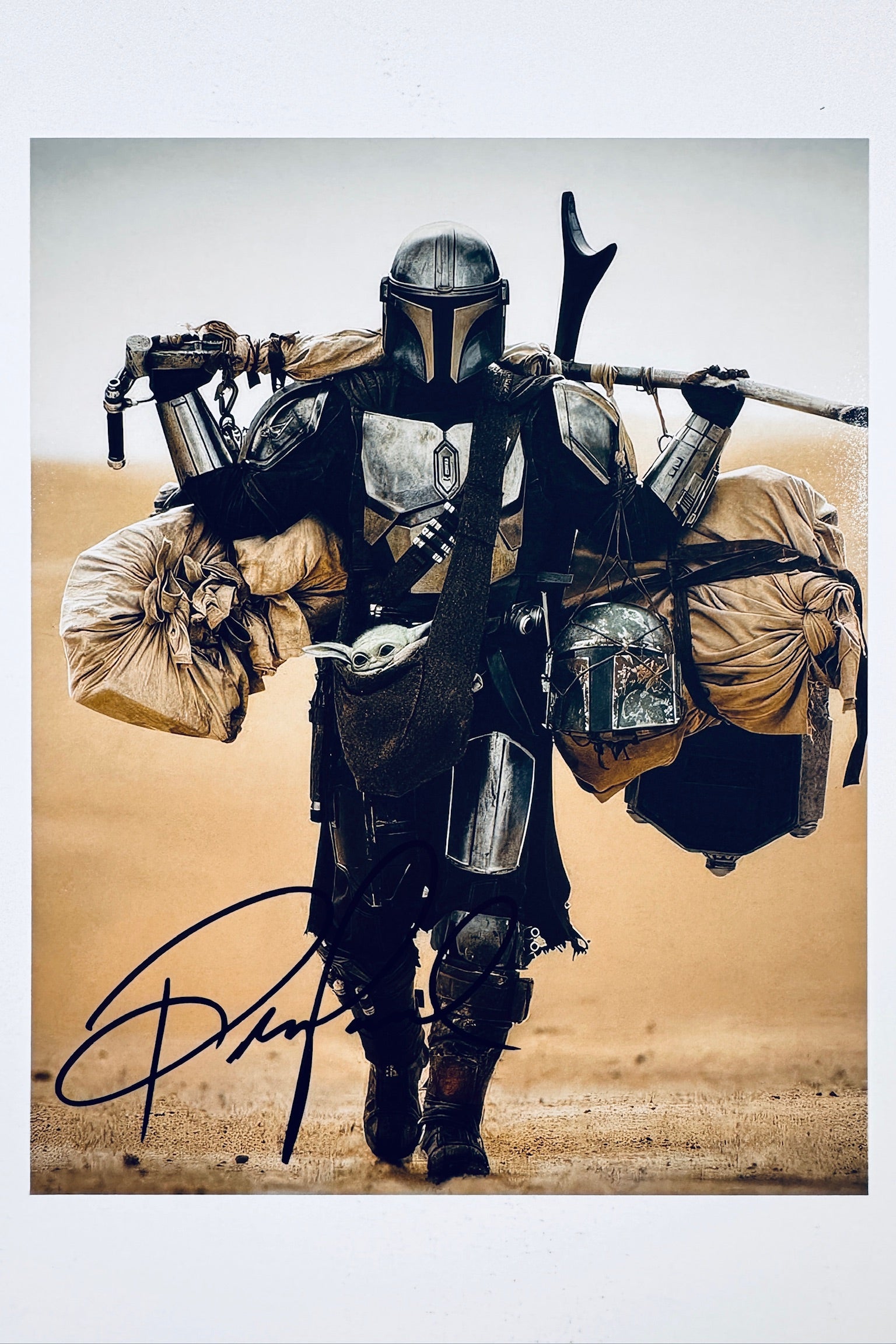 A promotional portrait of Pedro Pascal as the Mandalorian wearing beskar armor and walking through a desert carrying heavy bags and Grogu in a pouch, featuring a black marker signature across the lower foreground.