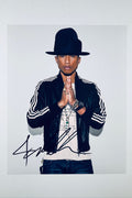 A portrait of Pharrell Williams wearing a large dark hat, black striped jacket, and layered necklaces with his hands held together in a prayer pose, featuring a dark marker signature across the bottom.