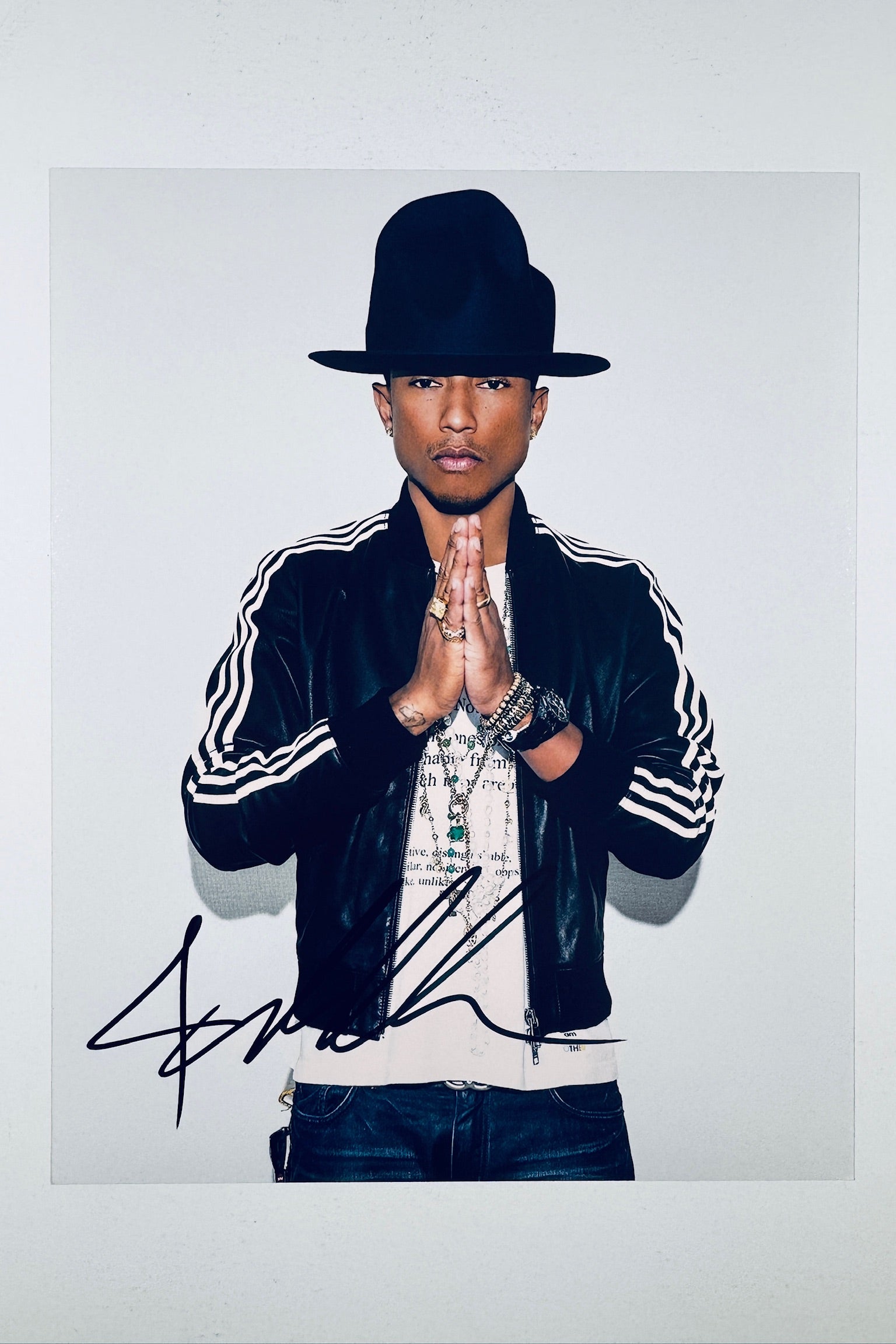 A portrait of Pharrell Williams wearing a large dark hat, black striped jacket, and layered necklaces with his hands held together in a prayer pose, featuring a dark marker signature across the bottom.