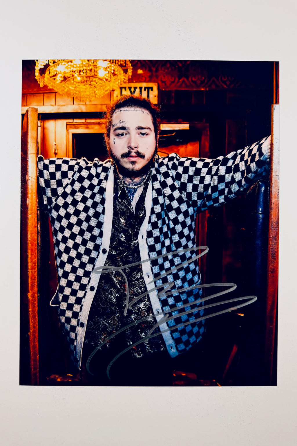 A portrait of Post Malone wearing a blue-and-white checkered cardigan and a patterned button-down shirt inside a dimly lit venue with a glowing exit sign and chandelier overhead, featuring a silver marker signature across the front of his cardigan.