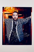 A portrait of Post Malone wearing a blue-and-white checkered cardigan and a patterned button-down shirt inside a dimly lit venue with a glowing exit sign and chandelier overhead, featuring a silver marker signature across the front of his cardigan.