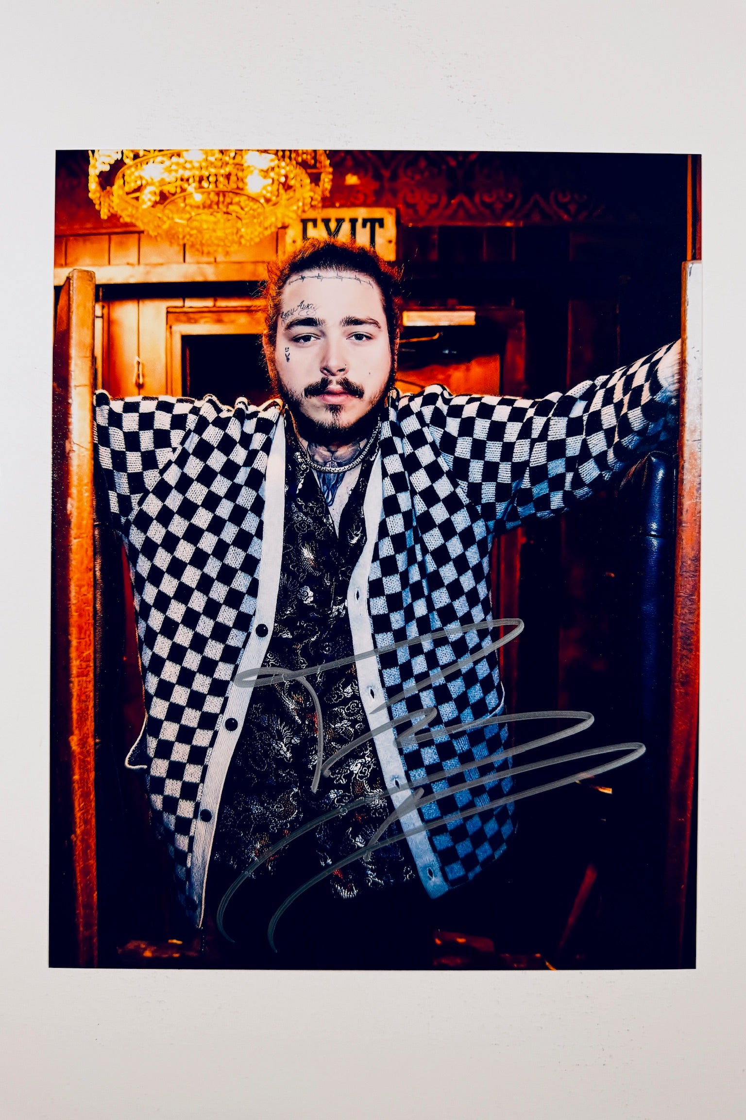 A portrait of Post Malone wearing a blue-and-white checkered cardigan and a patterned button-down shirt inside a dimly lit venue with a glowing exit sign and chandelier overhead, featuring a silver marker signature across the front of his cardigan.