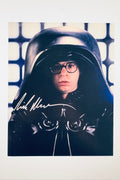 A portrait of Rick Moranis as Dark Helmet from Spaceballs wearing a large helmet and round glasses in front of a grid wall, featuring a silver marker signature across the lower left of the helmet.