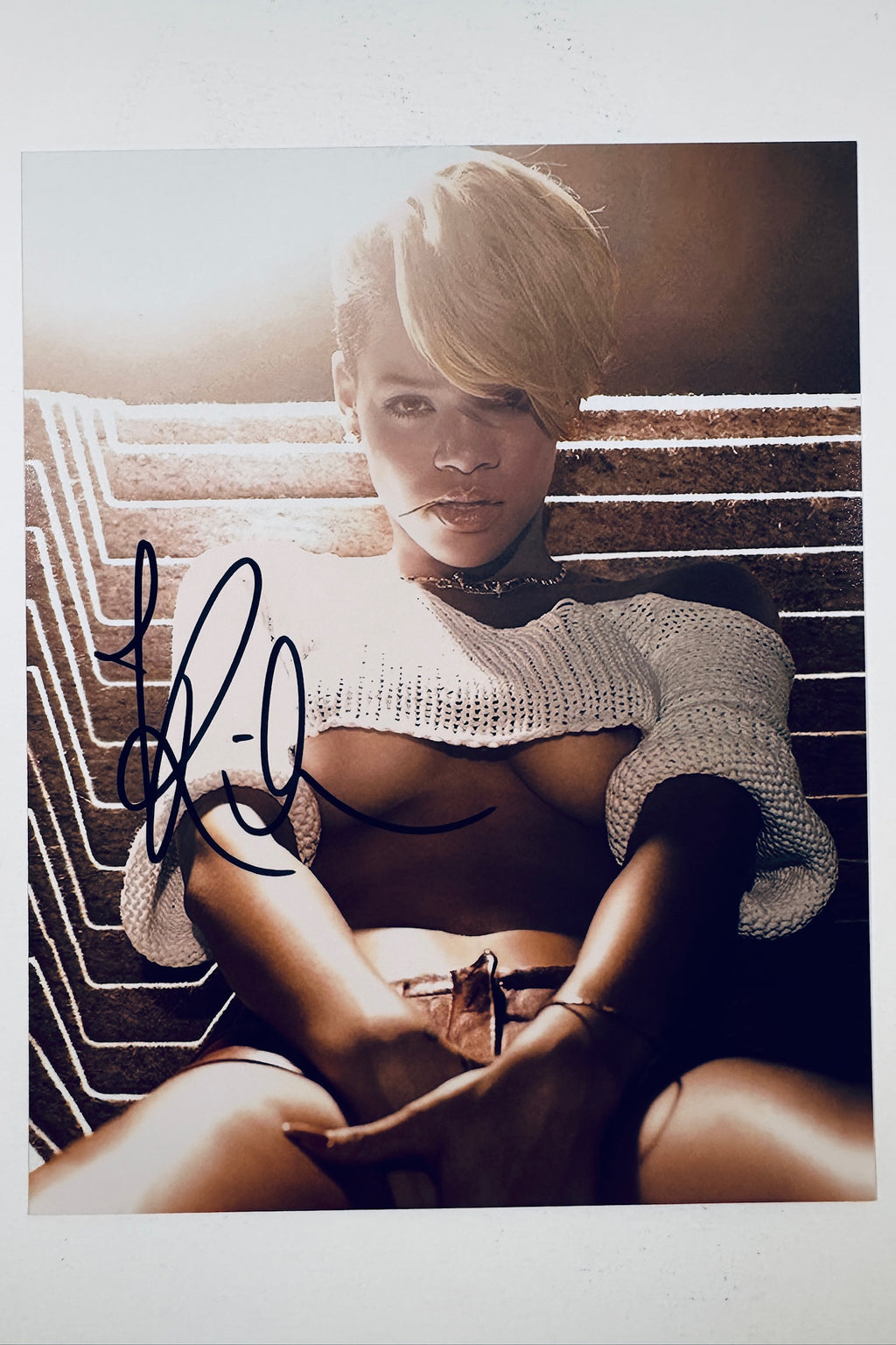A portrait of Rihanna with short blonde hair wearing a white crop top against a textured background, featuring a bold black marker signature across the left side of the image.