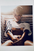 A portrait of Rihanna with short blonde hair wearing a white crop top against a textured background, featuring a bold black marker signature across the left side of the image.