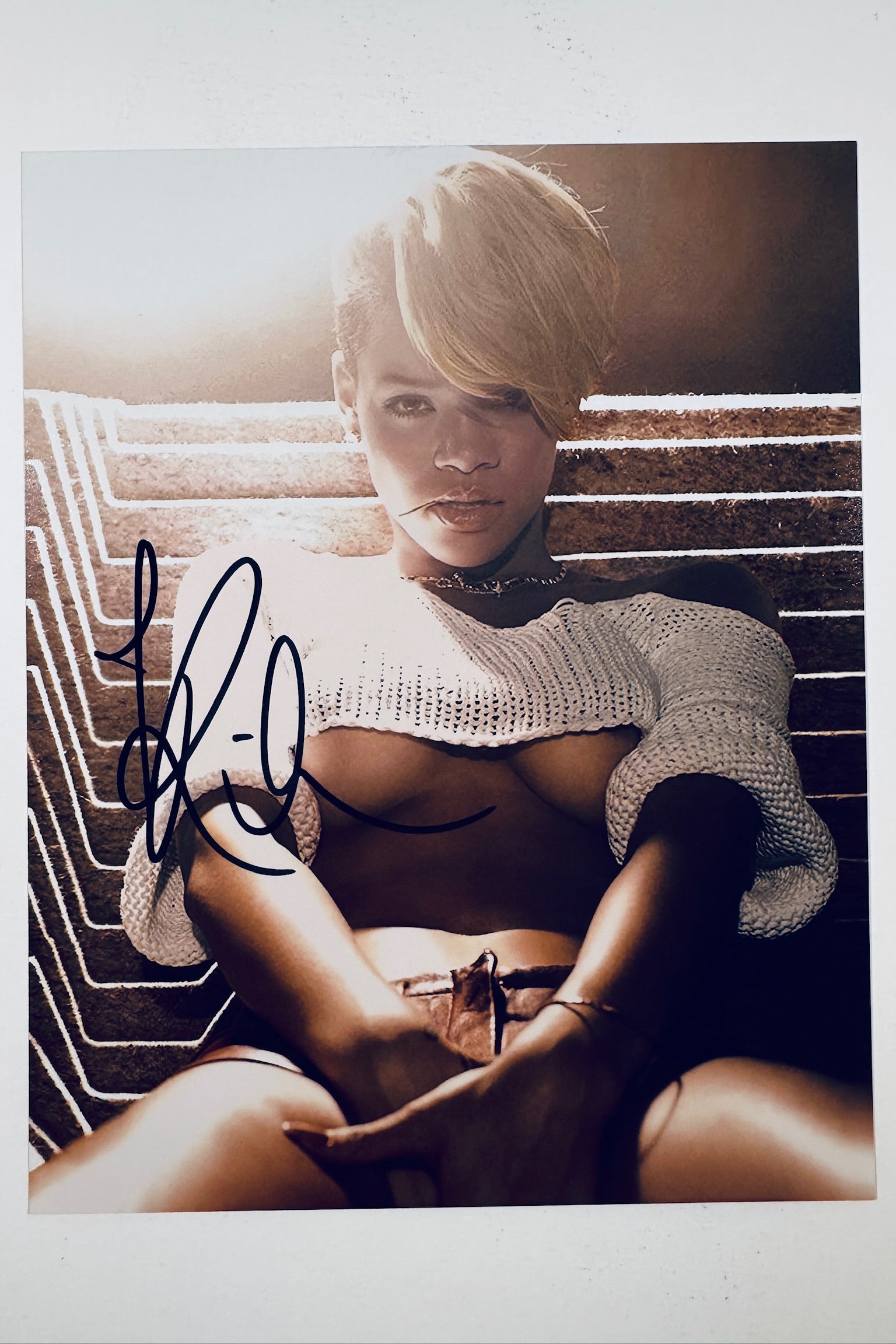 A portrait of Rihanna with short blonde hair wearing a white crop top against a textured background, featuring a bold black marker signature across the left side of the image.
