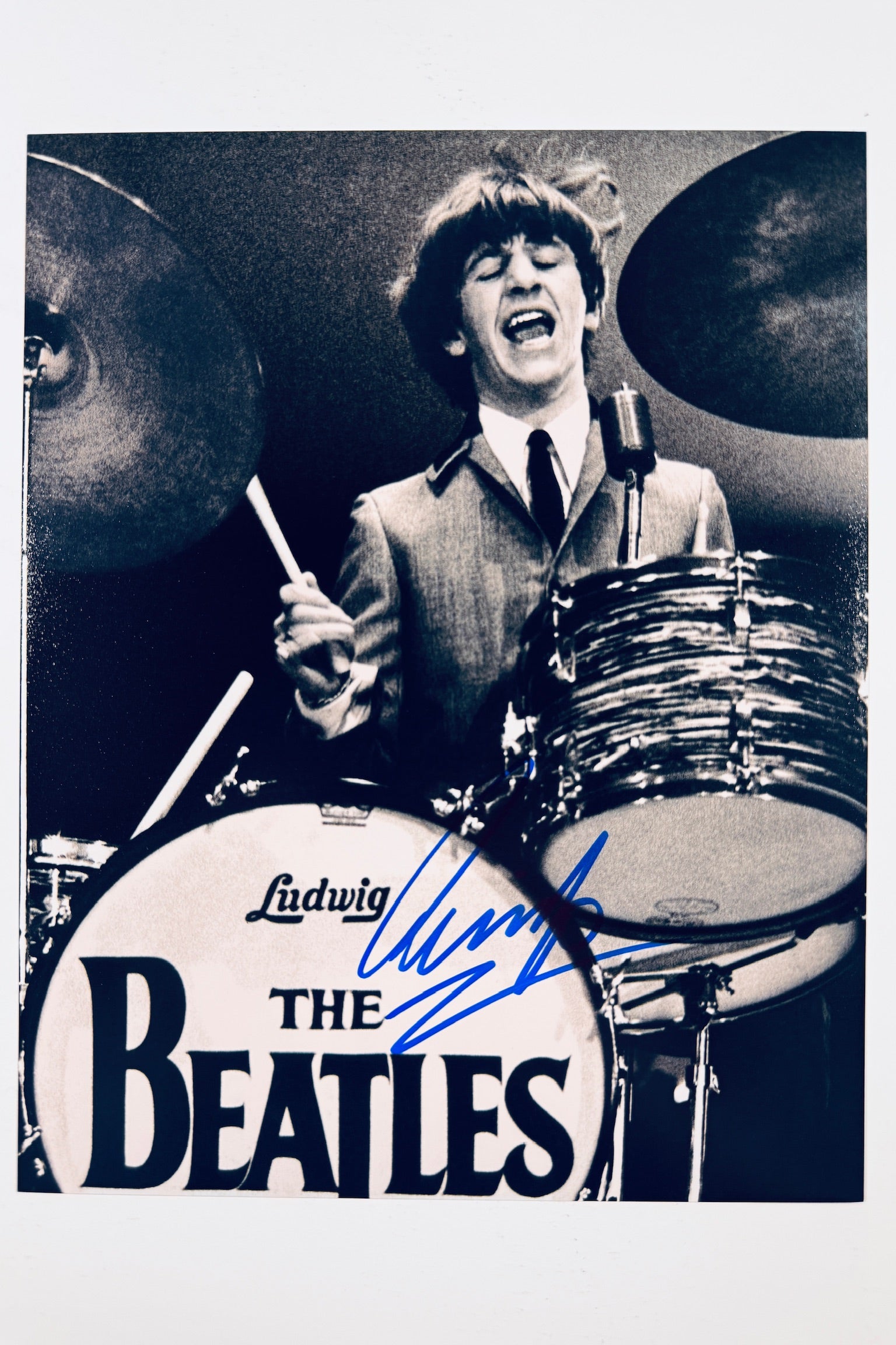 A black-and-white performance photograph of Ringo Starr playing drums behind a Ludwig "The Beatles" bass drum kit with cymbals, featuring a blue marker signature across the drum head.
