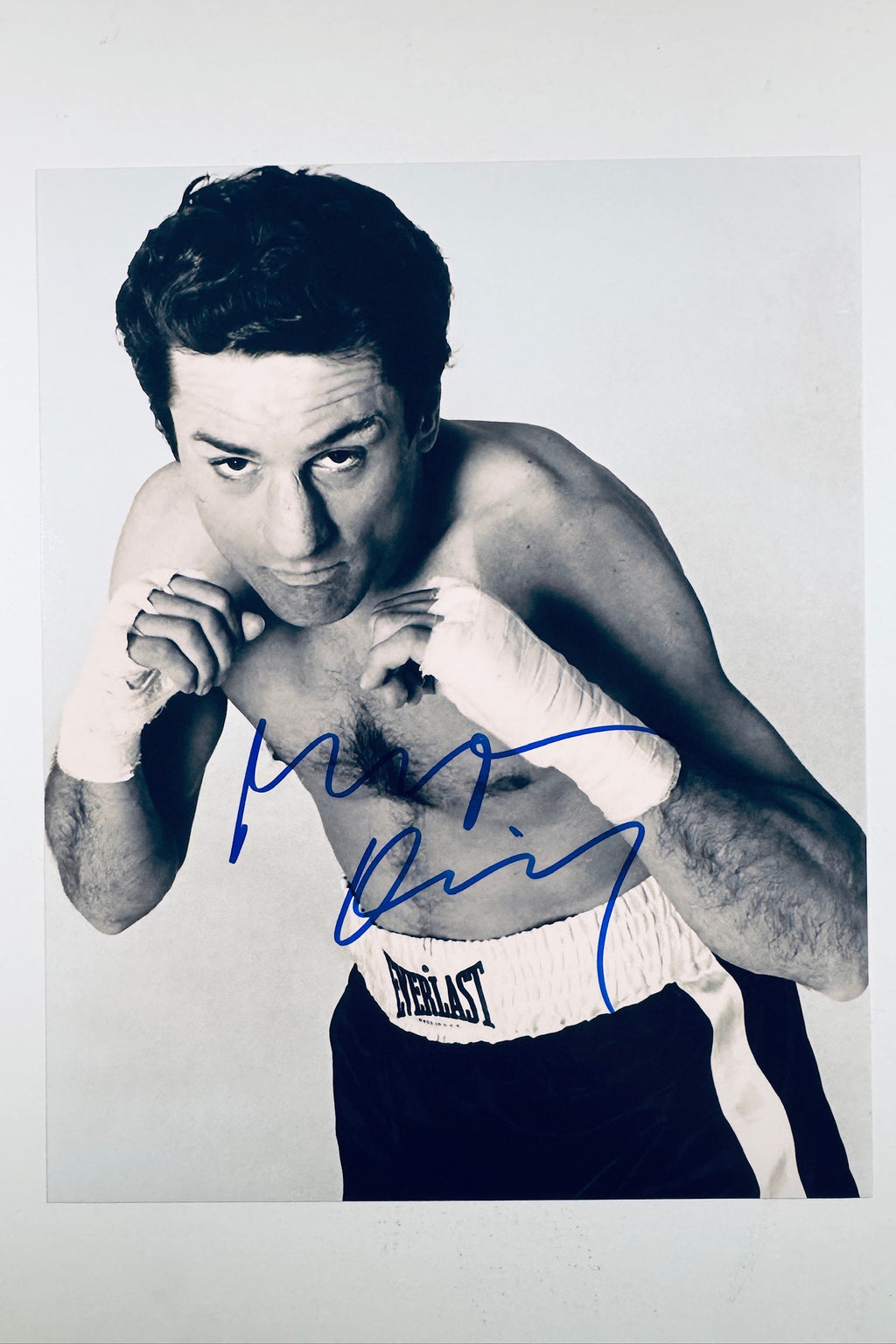 A black-and-white promotional portrait of Robert De Niro as Jake LaMotta in Raging Bull wearing Everlast boxing trunks and wrapped hands, featuring a blue autograph across his chest.