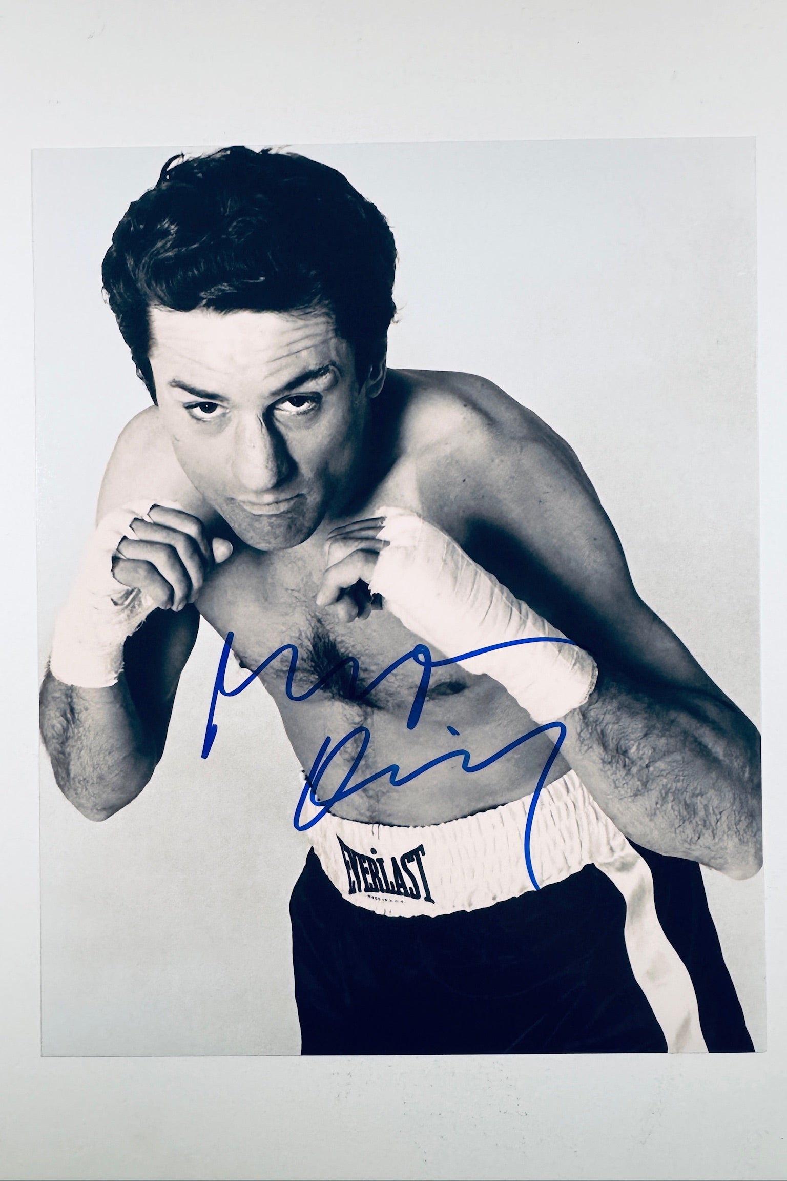 A black-and-white promotional portrait of Robert De Niro as Jake LaMotta in Raging Bull wearing Everlast boxing trunks and wrapped hands, featuring a blue autograph across his chest.
