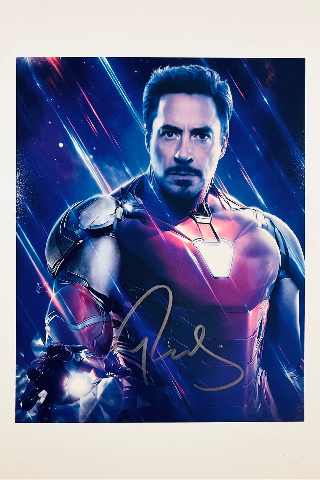 A portrait of Robert Downey Jr. in his Iron Man armor with a glowing hand repulsor against a cosmic blue and red background, featuring a gold marker signature across the lower center.