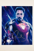 A portrait of Robert Downey Jr. in his Iron Man armor with a glowing hand repulsor against a cosmic blue and red background, featuring a gold marker signature across the lower center.