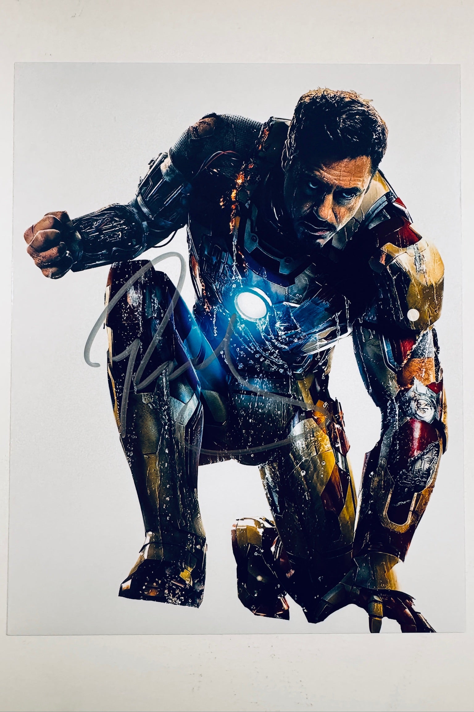 A promotional photo of Robert Downey Jr. as Iron Man in a battle-damaged suit crouching with a glowing arc reactor, featuring an autograph across the right leg area.