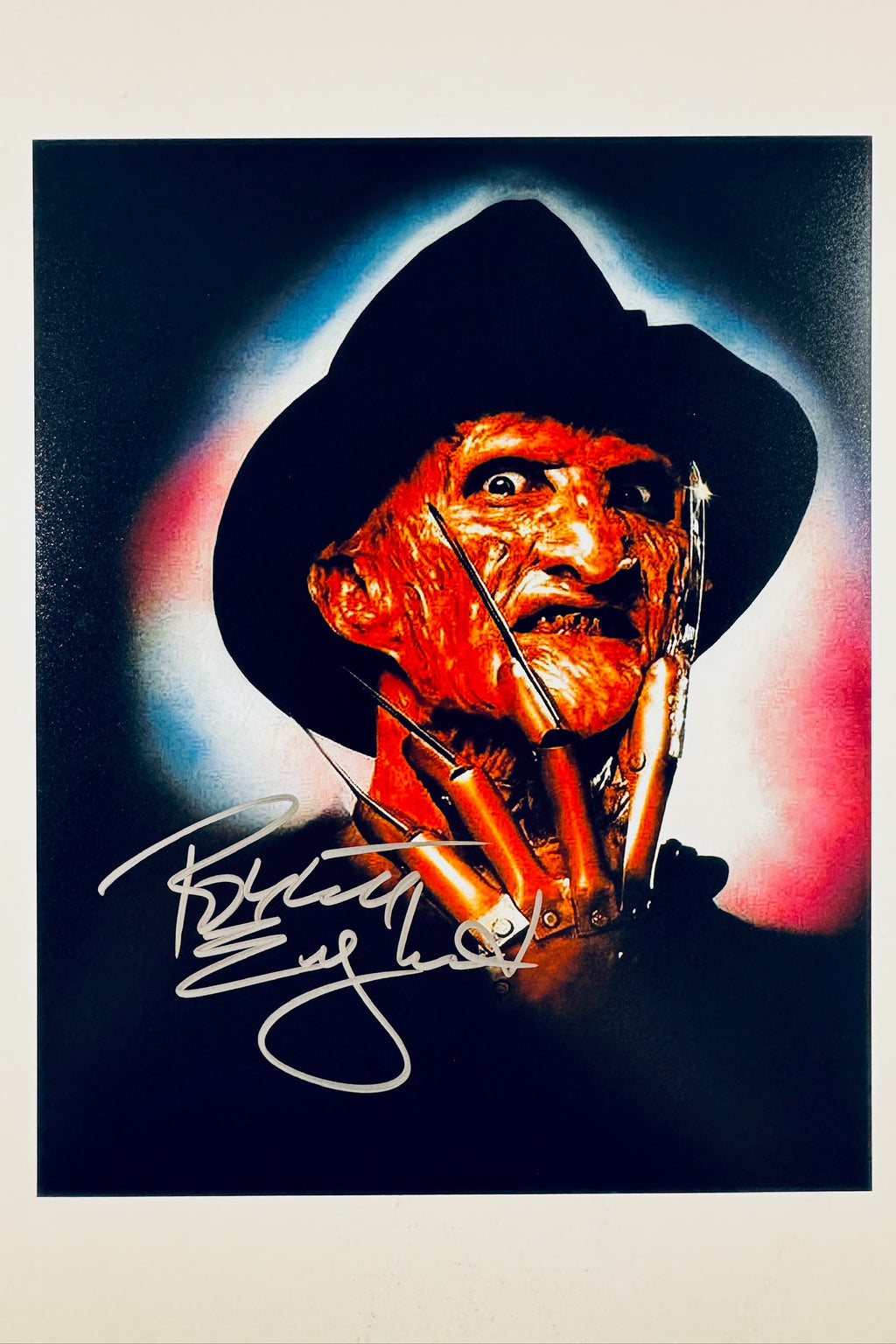 A promotional portrait of Robert Englund as Freddy Krueger in A Nightmare on Elm Street wearing his signature striped sweater, fedora, and bladed glove, featuring a light autograph signed across the lower portion.
