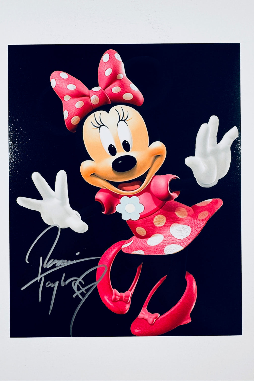 A portrait of Minnie Mouse against a black background posing with her arms out and wearing a red polka-dot dress and bow, featuring a silver marker signature across the lower left.