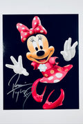 A portrait of Minnie Mouse against a black background posing with her arms out and wearing a red polka-dot dress and bow, featuring a silver marker signature across the lower left.