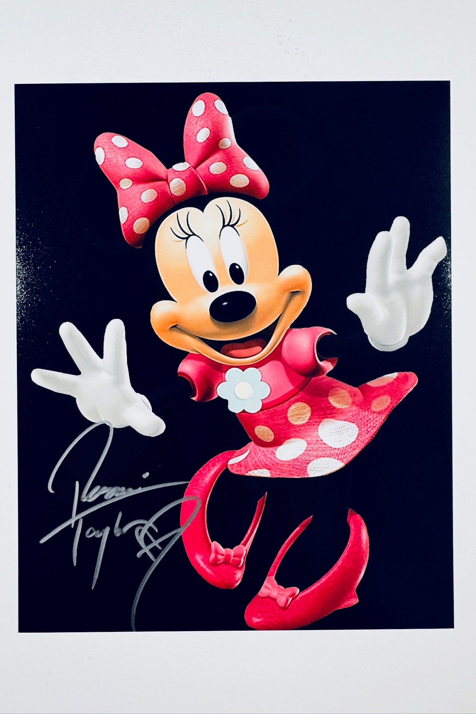 A portrait of Minnie Mouse against a black background posing with her arms out and wearing a red polka-dot dress and bow, featuring a silver marker signature across the lower left.