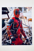 A promotional portrait of Ryan Reynolds as Deadpool in his red and black tactical suit holding a dagger with a chaotic background, featuring a silver marker signature across his chest.