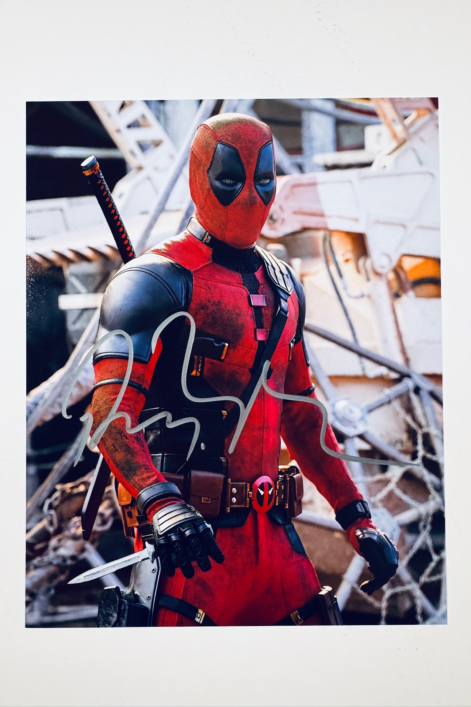 A promotional portrait of Ryan Reynolds as Deadpool in his red and black tactical suit holding a dagger with a chaotic background, featuring a silver marker signature across his chest.
