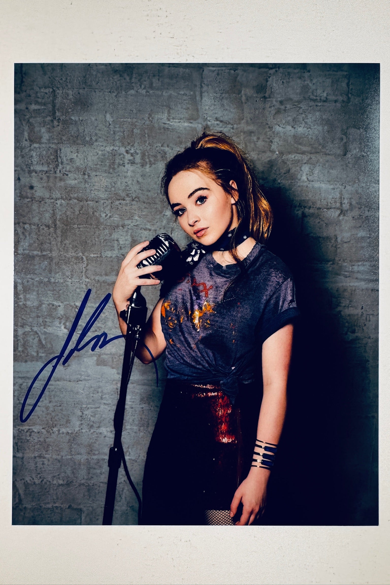 Signed full photo of Sabrina Carpenter holding a vintage microphone against a brick wall backdrop, featuring a blue autograph.