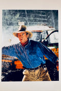 A portrait of Sam Neill as Dr. Alan Grant in a hat and blue shirt gesturing in the rain with a signature on the lower left.