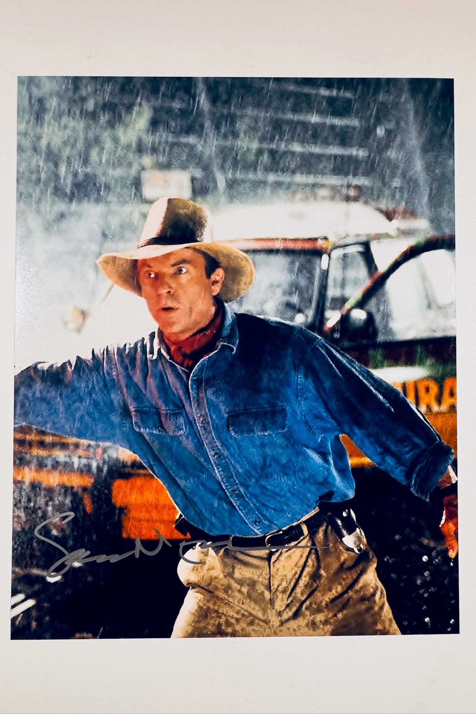 A portrait of Sam Neill as Dr. Alan Grant in a hat and blue shirt gesturing in the rain with a signature on the lower left.