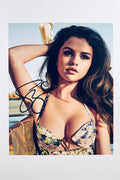 A portrait of Selena Gomez with dark wavy hair and dramatic cat-eye makeup wearing a patterned top outdoors with a bright sky, featuring a black marker signature across her right arm.