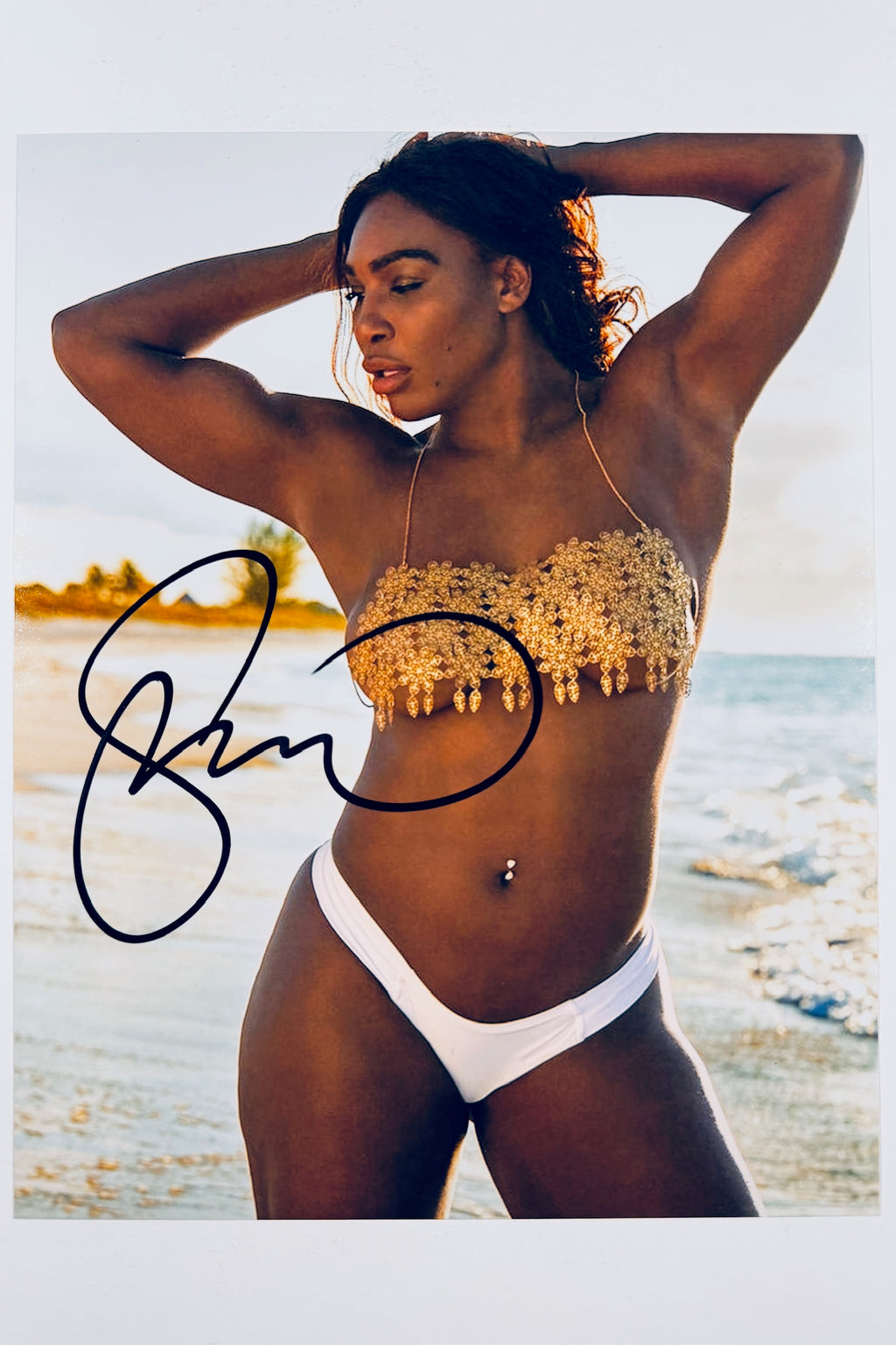 A portrait of Serena Williams on a beach wearing a gold-accented bikini top and white bottoms with her hands behind her head, featuring a dark marker signature across the left side.