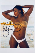 A portrait of Serena Williams on a beach wearing a gold-accented bikini top and white bottoms with her hands behind her head, featuring a dark marker signature across the left side.