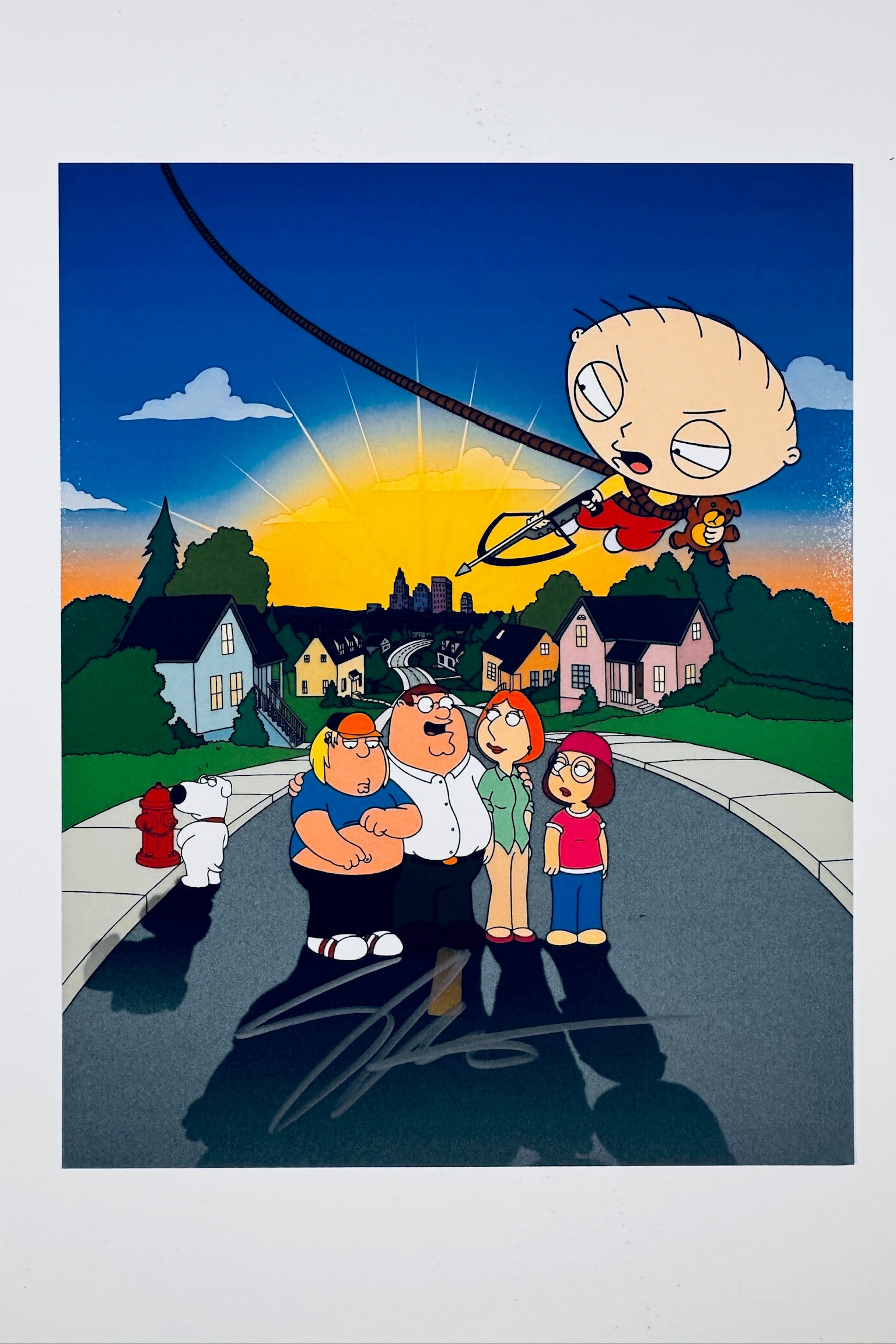 An illustrated art print of the Griffin family standing on a suburban street with Stewie flying overhead on a wire, featuring a silver marker signature across the pavement in the foreground.