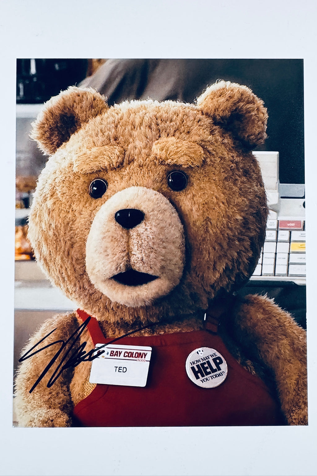 A portrait of the character Ted wearing a Bay Colony name tag and a "HOW MAY WE HELP YOU TODAY?" button, featuring a dark signature on the lower left.