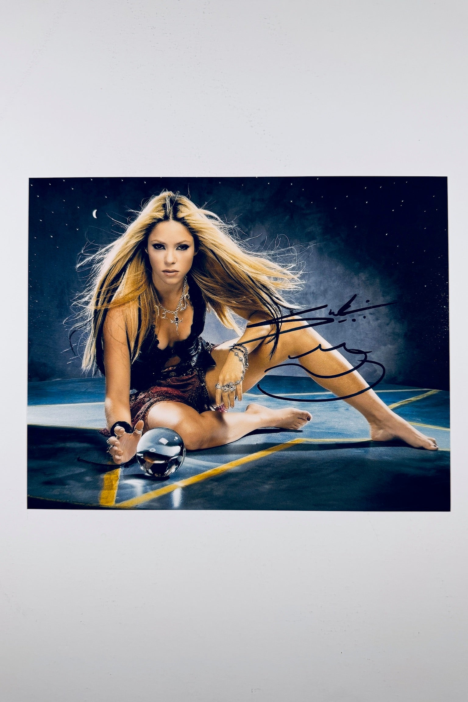 A portrait of Shakira sitting on the floor with long blonde hair and dark streaks, reaching toward a glass sphere on a surface, featuring a black marker signature across her right leg and hip.
