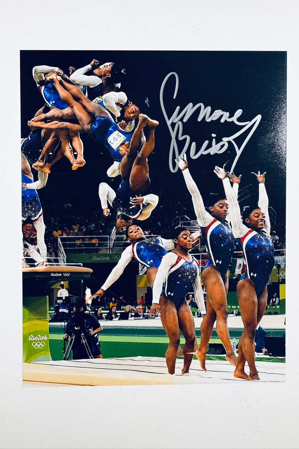 A composite action photograph of Simone Biles competing in a blue and red leotard at the Olympics, showing multiple phases of her vault routine and standing poses, featuring a silver marker signature in the upper right.