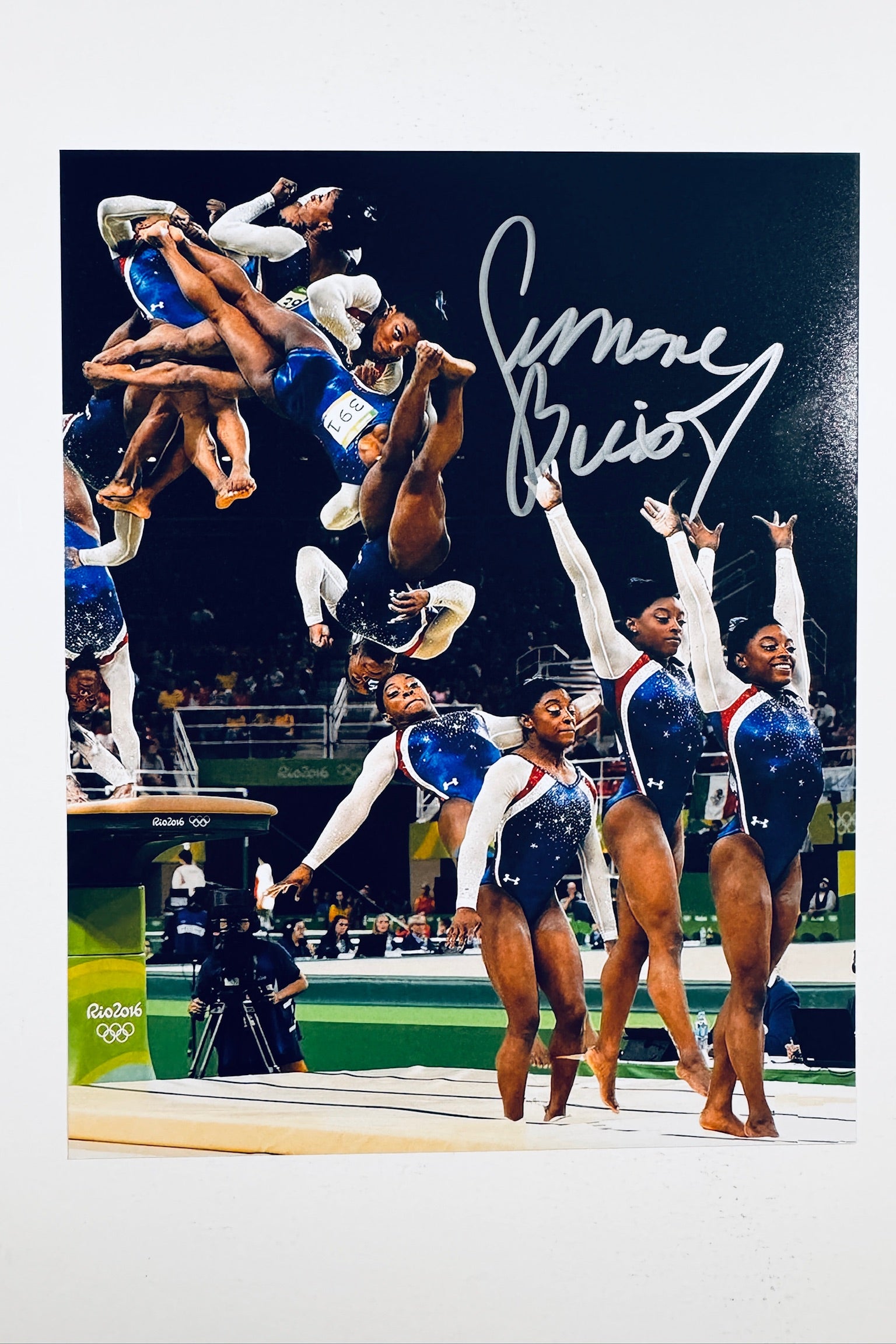 A composite action photograph of Simone Biles competing in a blue and red leotard at the Olympics, showing multiple phases of her vault routine and standing poses, featuring a silver marker signature in the upper right.