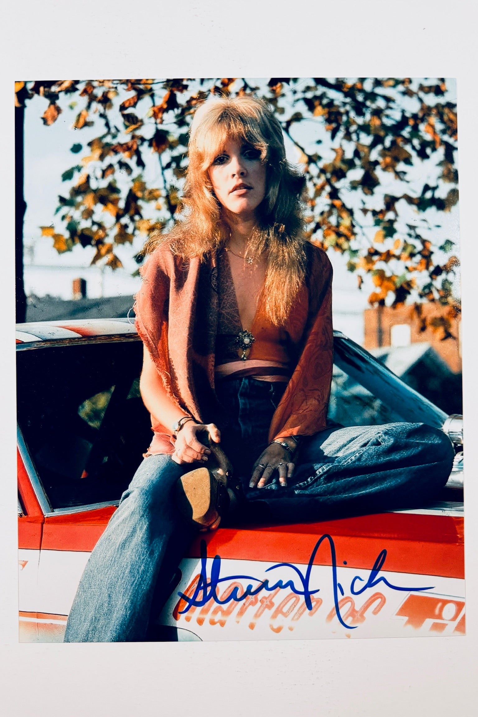 A portrait of Stevie Nicks sitting on the hood of a red and white car outdoors under autumn leaves, wearing a patterned top, shawl, and jeans, featuring a blue marker signature across the lower bumper.