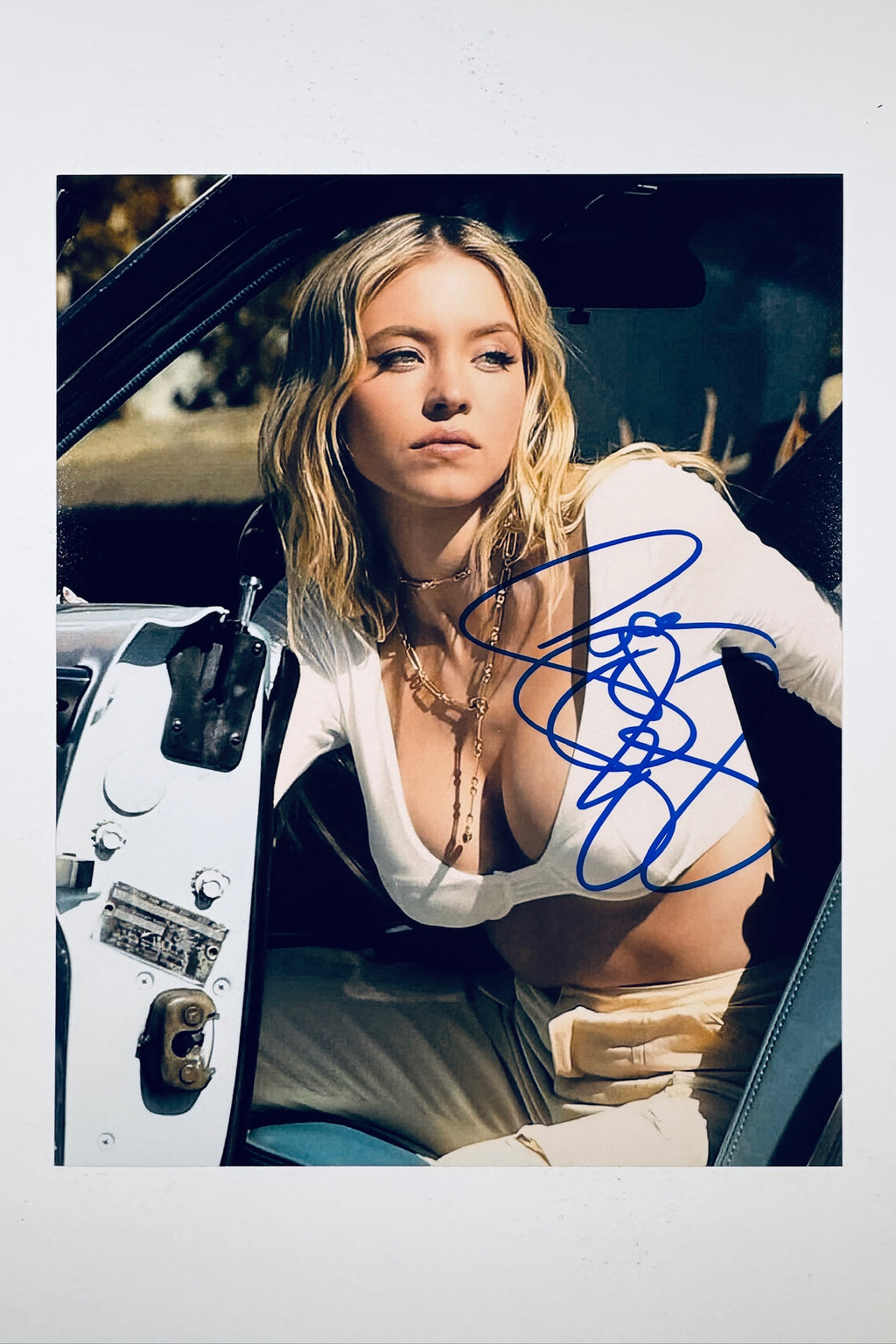 A portrait of Sydney Sweeney with blonde hair wearing a white top and layered necklaces while sitting inside a classic car with the door open, featuring a blue marker signature across her chest and torso.