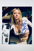 A portrait of Sydney Sweeney with blonde hair wearing a white top and layered necklaces while sitting inside a classic car with the door open, featuring a blue marker signature across her chest and torso.
