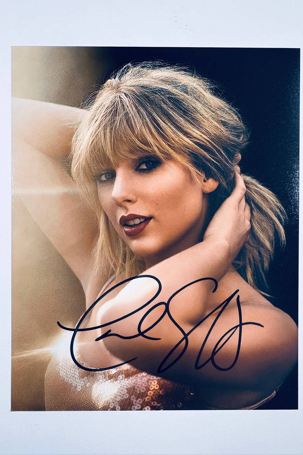 A portrait of Taylor Swift looking toward the camera with her hands near her hair and neck, wearing a light sequined top, featuring a black marker signature across her lower shoulder and chest.