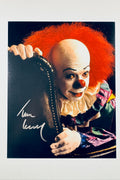A promotional portrait of Tim Curry as Pennywise the Clown from Stephen King's It, featuring vibrant red hair, white face paint, a red nose, and a colorful collar, with a white autograph signed across the lower left side.