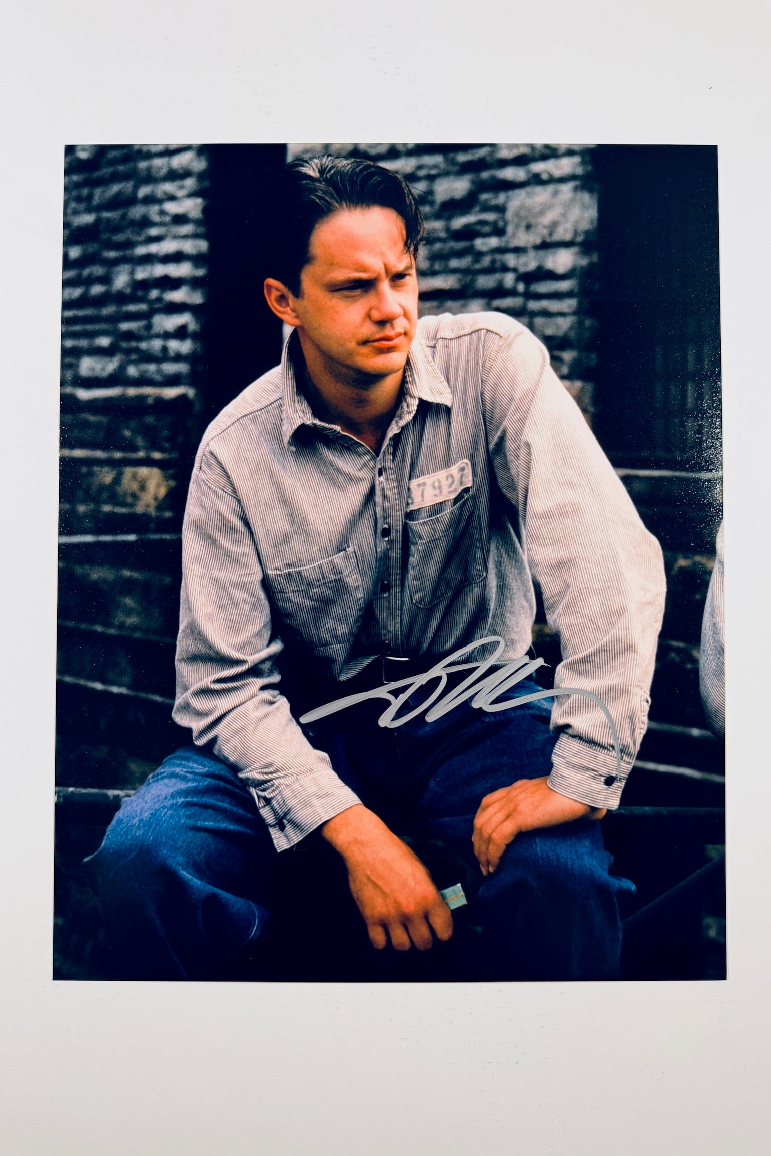 A promotional portrait of Tim Robbins as Andy Dufresne wearing a striped shirt and jeans seated against a stone wall, featuring a silver marker signature across his lap.