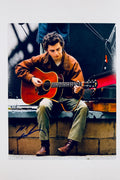 A portrait of Timothée Chalamet sitting on a ledge outdoors playing an acoustic guitar while wearing a brown jacket and green pants, featuring a dark marker signature across the lower left.