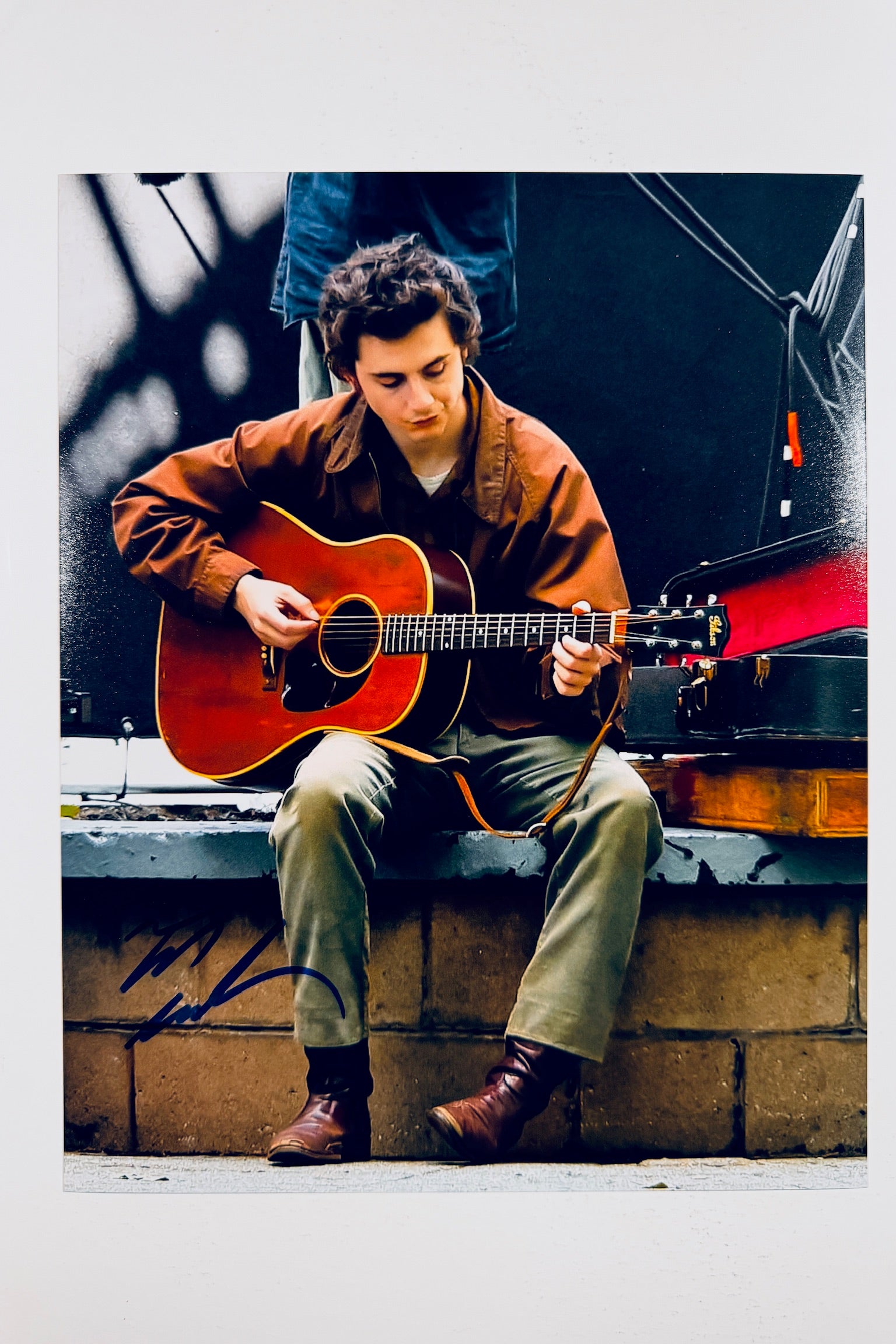 A portrait of Timothée Chalamet sitting on a ledge outdoors playing an acoustic guitar while wearing a brown jacket and green pants, featuring a dark marker signature across the lower left.