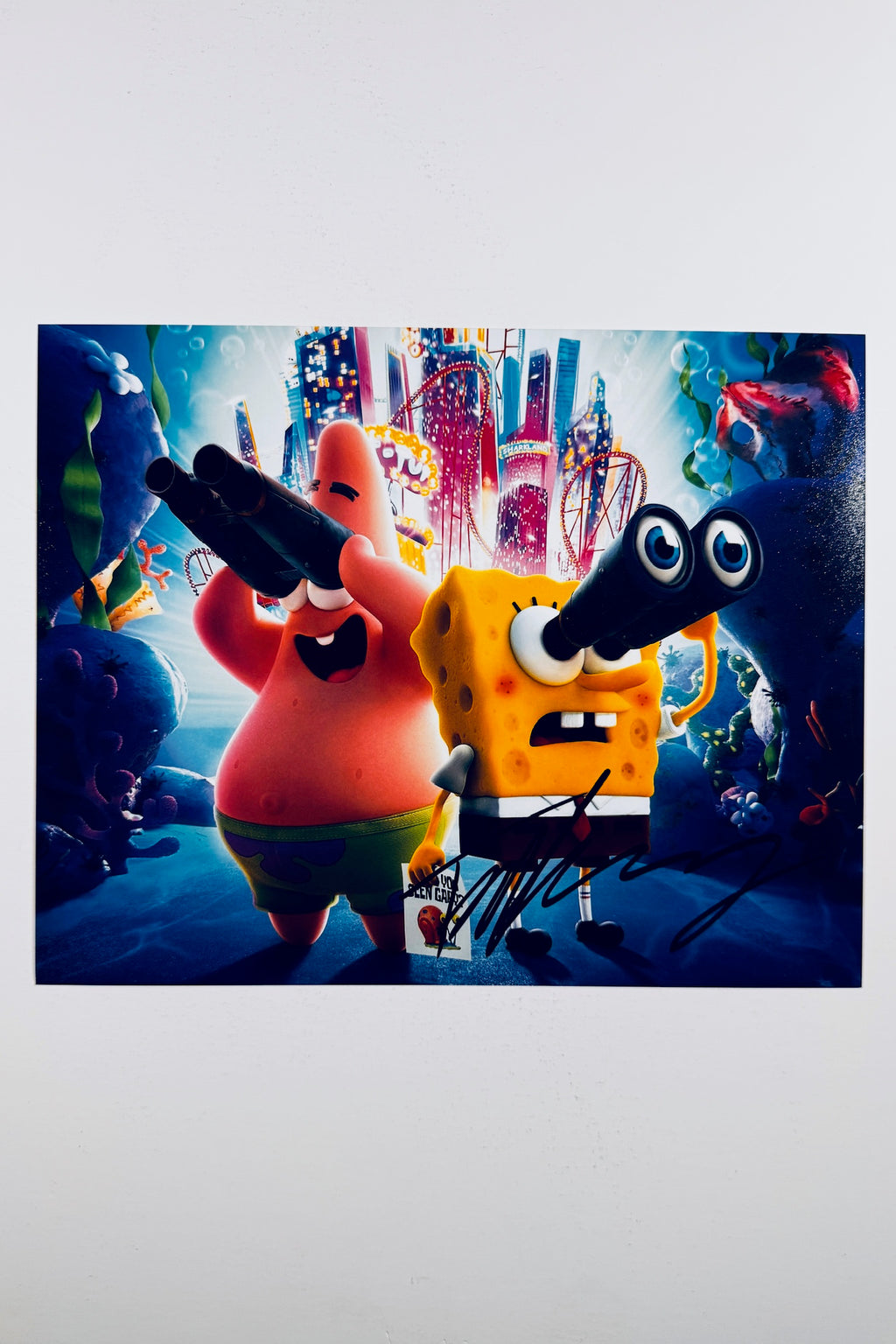 A vibrant animated scene of SpongeBob SquarePants and Patrick Star looking through binoculars underwater against a brightly lit city skyline background, featuring a bold black marker signature across the lower right foreground.