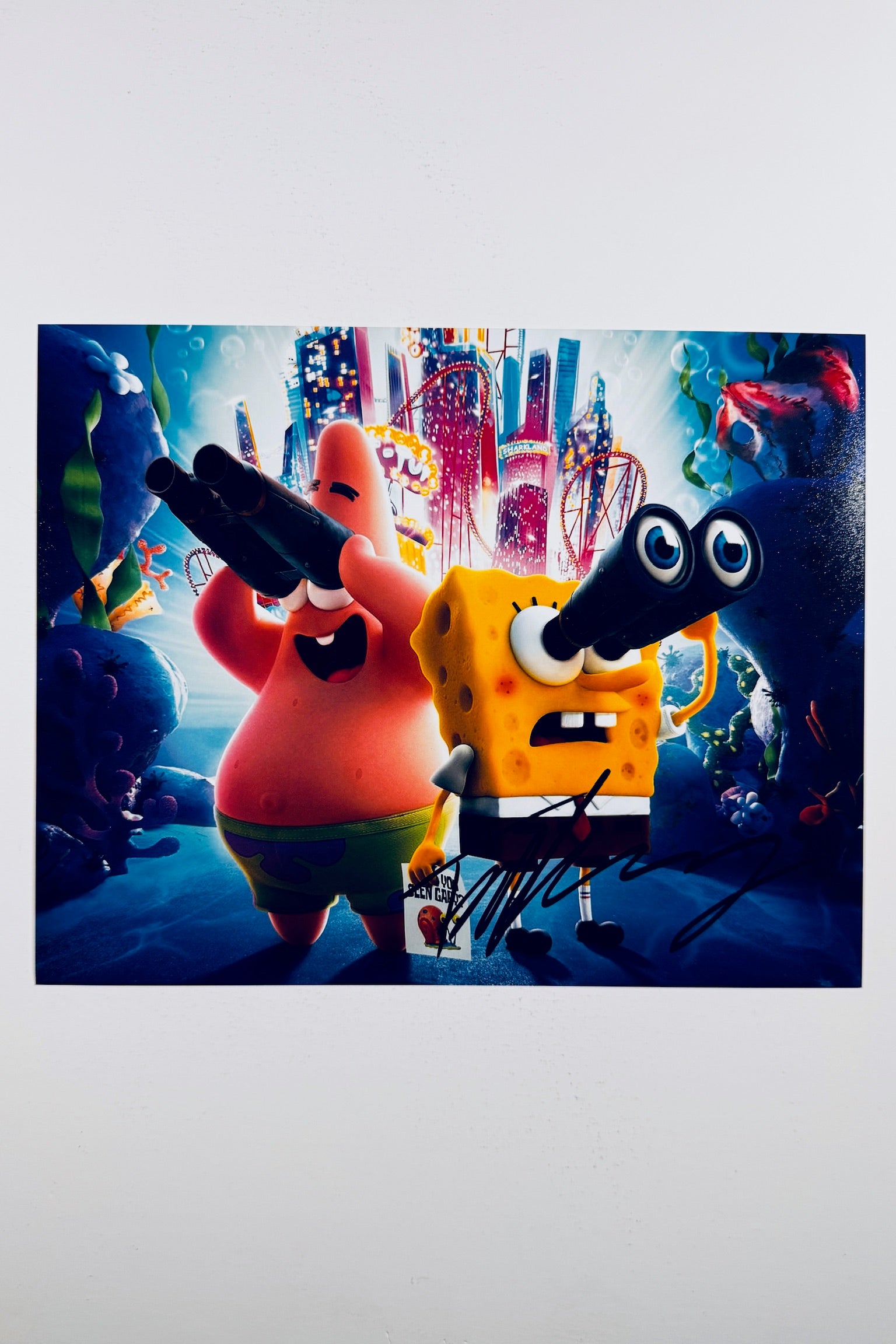 A vibrant animated scene of SpongeBob SquarePants and Patrick Star looking through binoculars underwater against a brightly lit city skyline background, featuring a bold black marker signature across the lower right foreground.