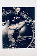 A black-and-white performance shot of Travis Scott mid-air with his leg kicked out on stage, featuring a silver marker signature across the lower right.