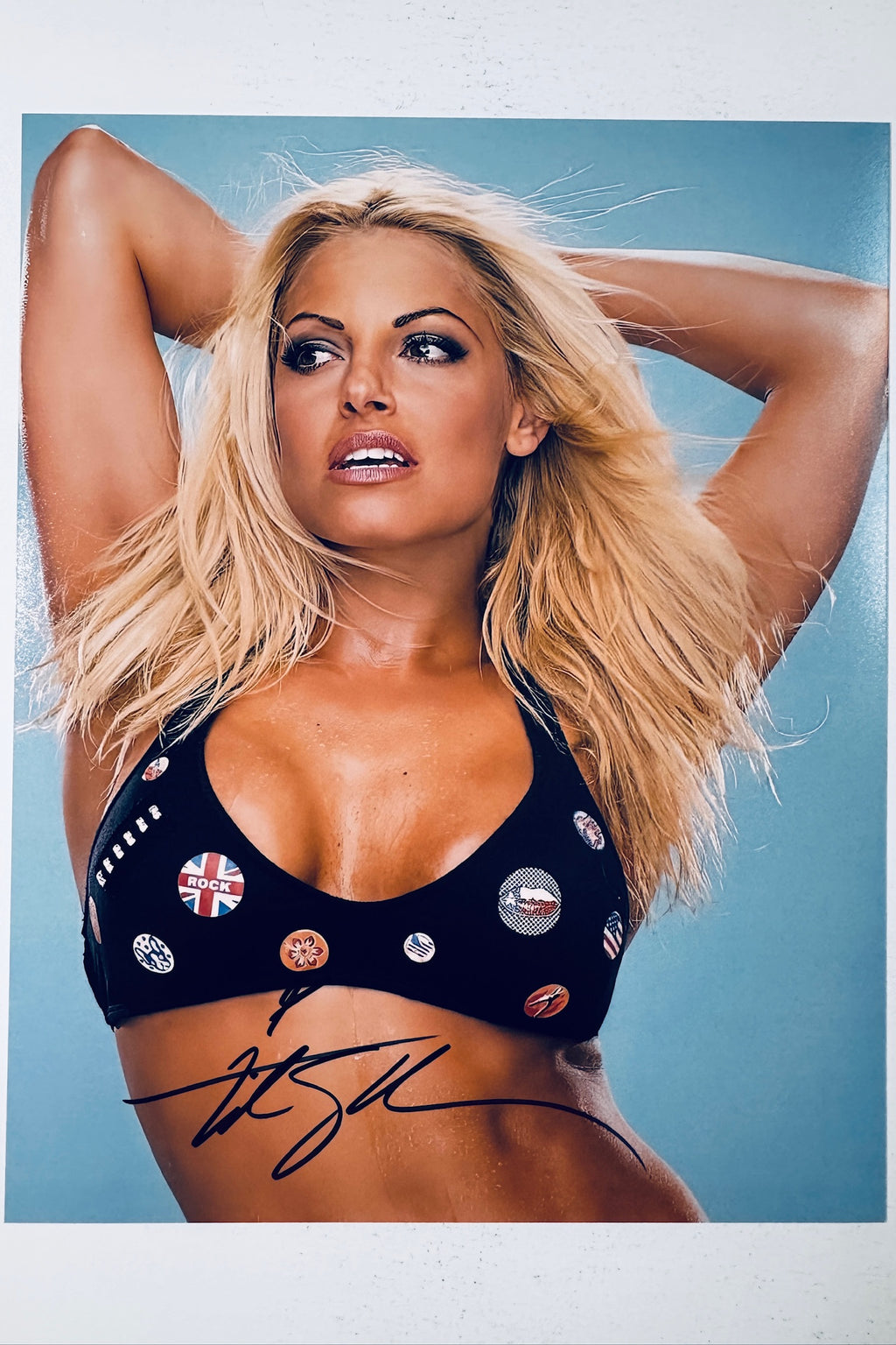 Signed 8x10 matte promotional photo of WWE Hall of Famer Trish Stratus posing in a black bikini top with buttons against a light blue background, featuring a bold black ink autograph.