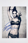 A black and white studio portrait of Victoria Beckham posing in lace underwear against a textured wall with her arms raised, featuring a blue marker signature across the left side.